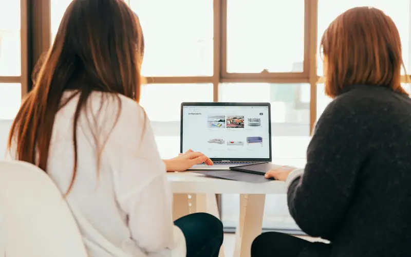 Two women sit side by side at a white table by a large window, looking at a laptop together