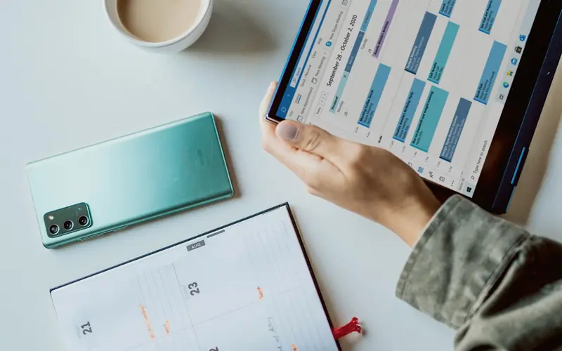 A planner, a phone and a tablet showing a colour-coded schedule on a white desk beside a cup of coffee